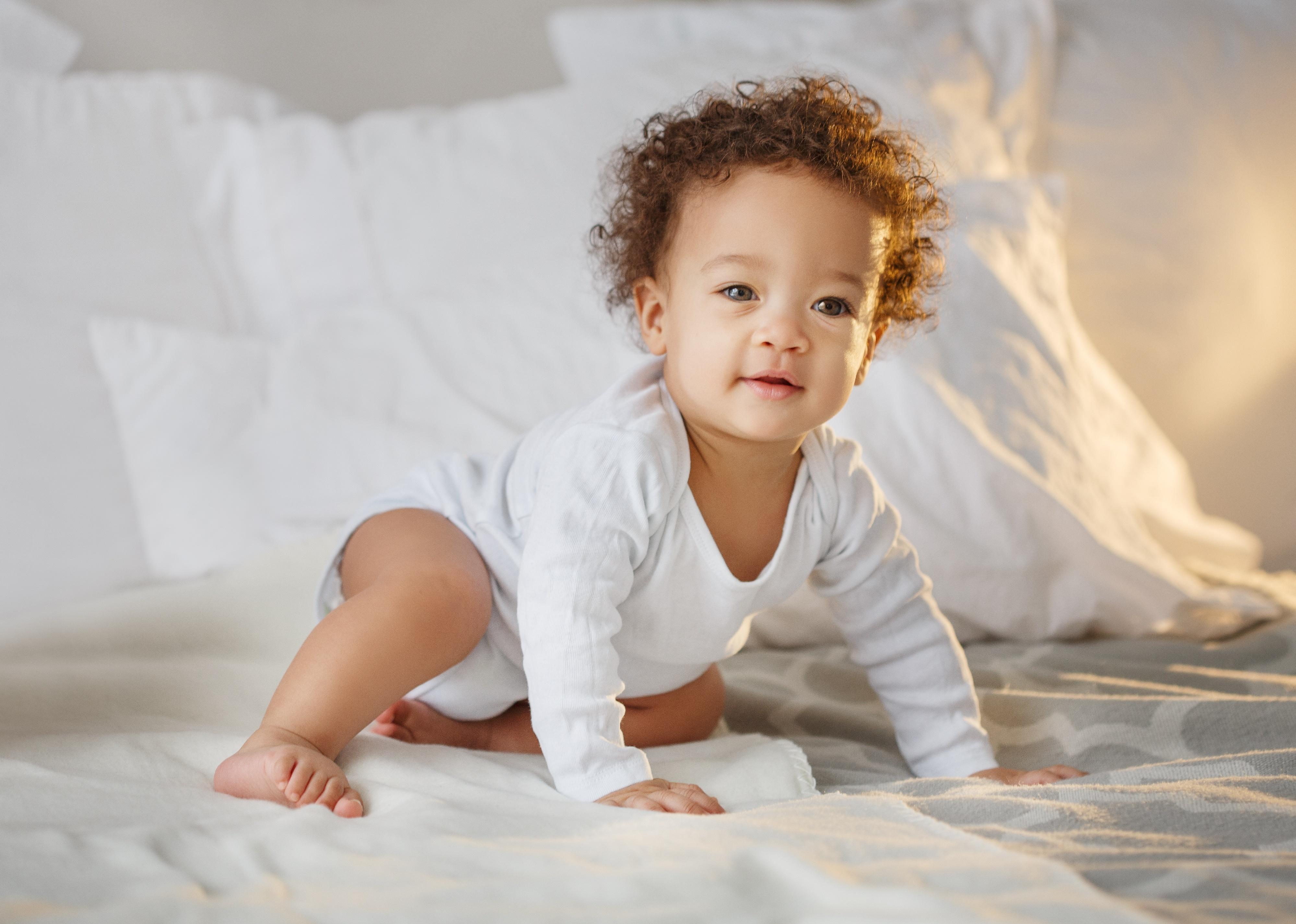 Baby with curly hair sitting up in bed.