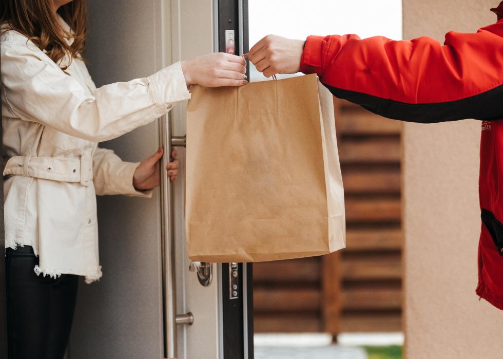 A food delivery person passes a brown paper bag to a customer at the door.