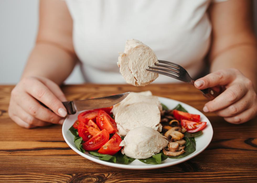 A woman eating baked chicken breasts with greens, tomatoes, and mushrooms.