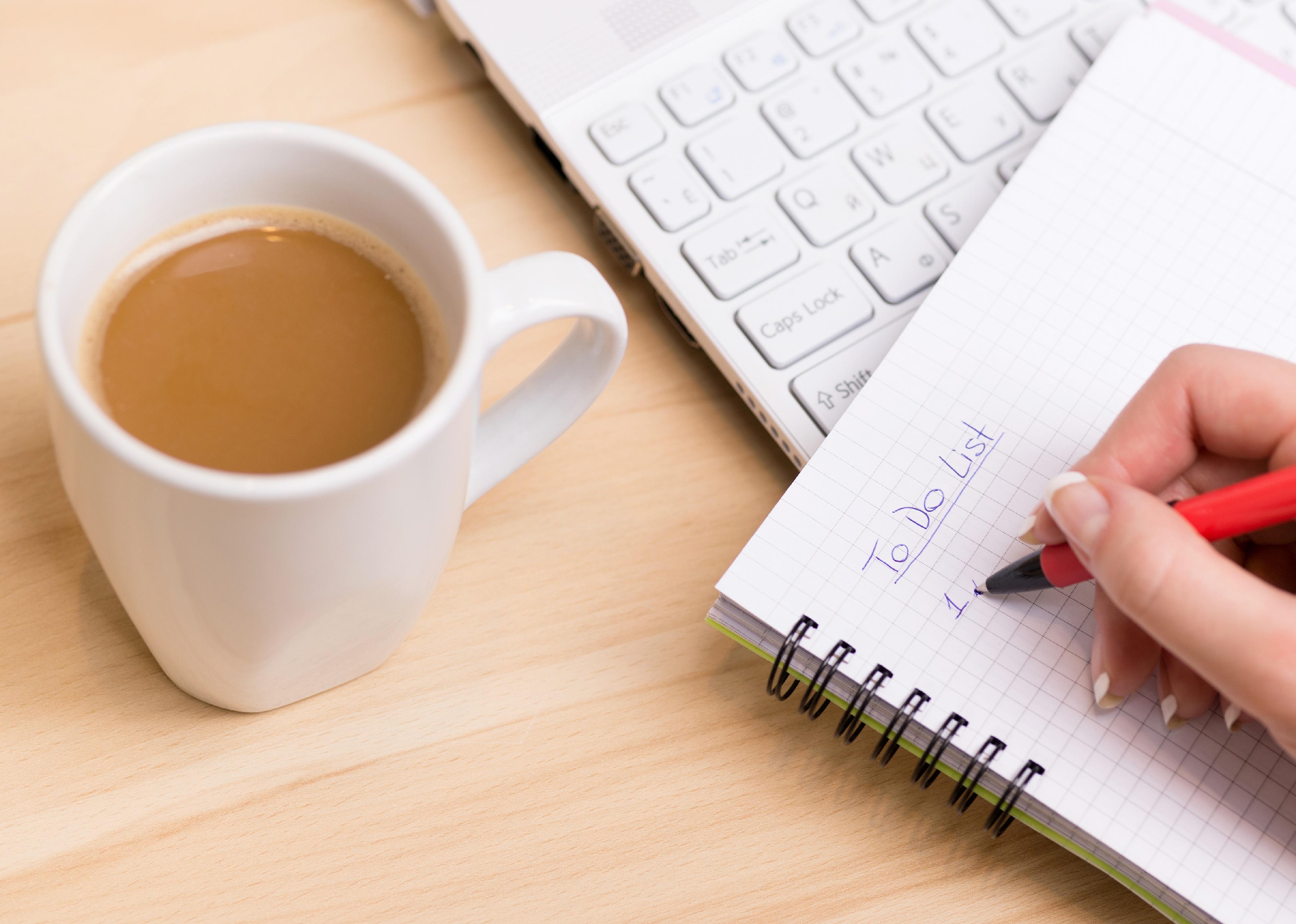 Person's hand writing a to-do list at their desk with a cup of fresh coffee.