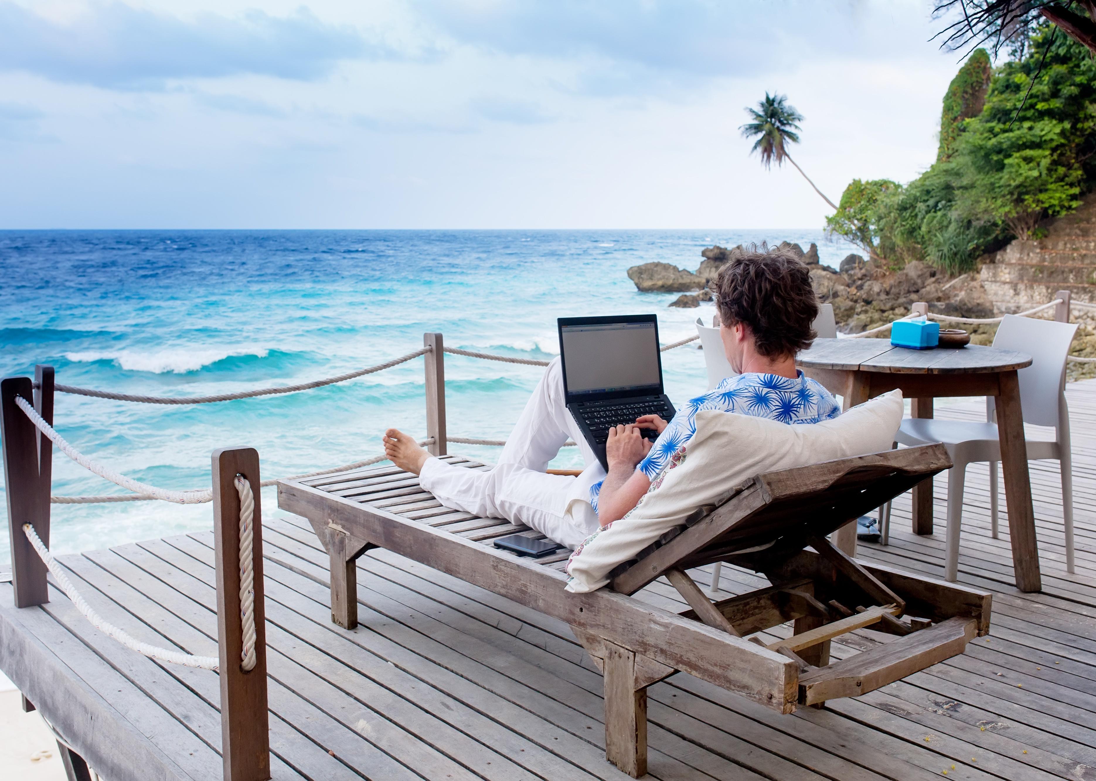 Person with laptop and mobile phone working remotely on a lounge chair overlooking the ocean. 