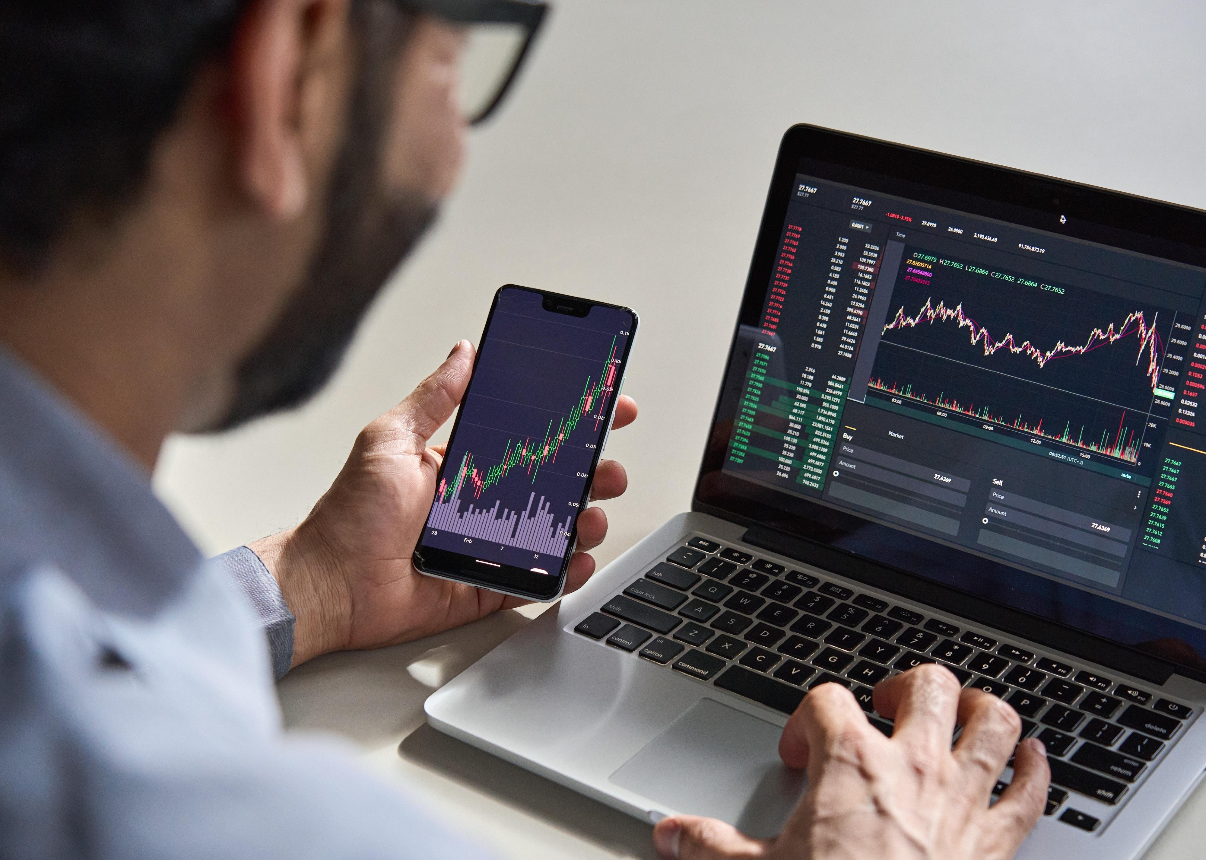 Man with trading data index chart graph on smartphone and laptop screen.