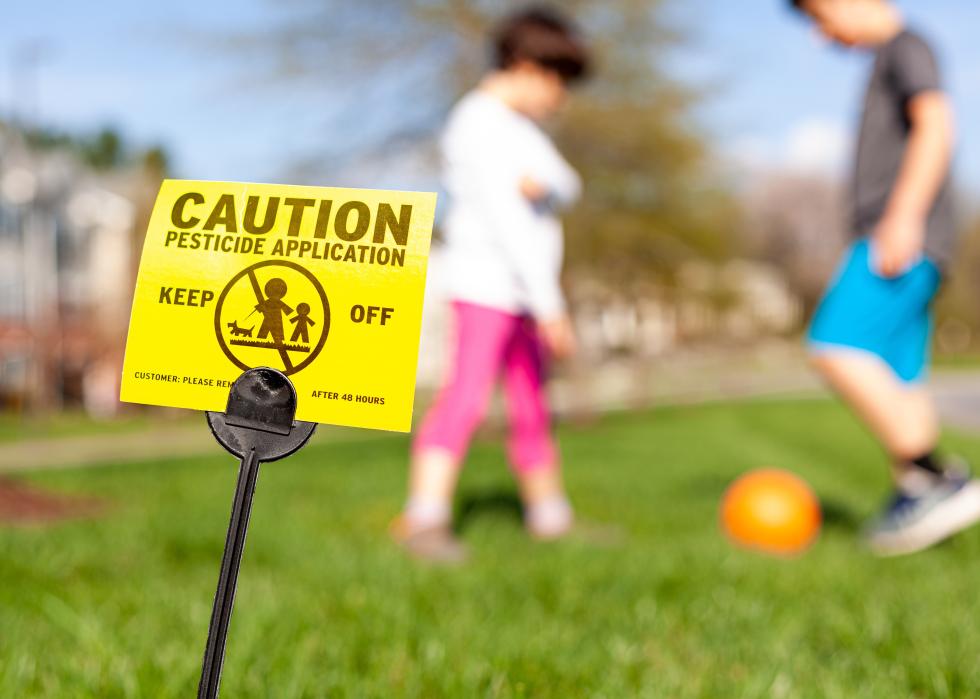 A yellow sign stuck in a lawn that says, caution, pesticide applications, keep off, and has a symbol showing an adult and a child walking a dog on a leash. In the blurred background, kids play with a ball. 
