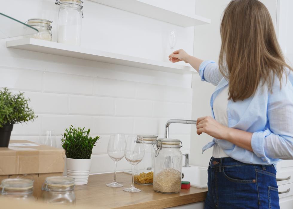 A woman places the first clean wine glass on an empty shelf, with other jars and glasses on the counter beside her.