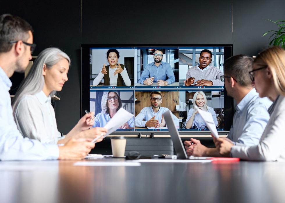 Videoconference in meeting room with people sitting in office and colleagues on monitor.