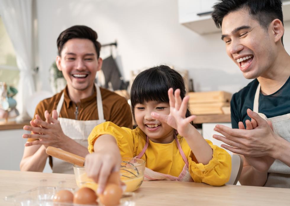 Two Asian men clapping and watching little Asian girl cracking eggs