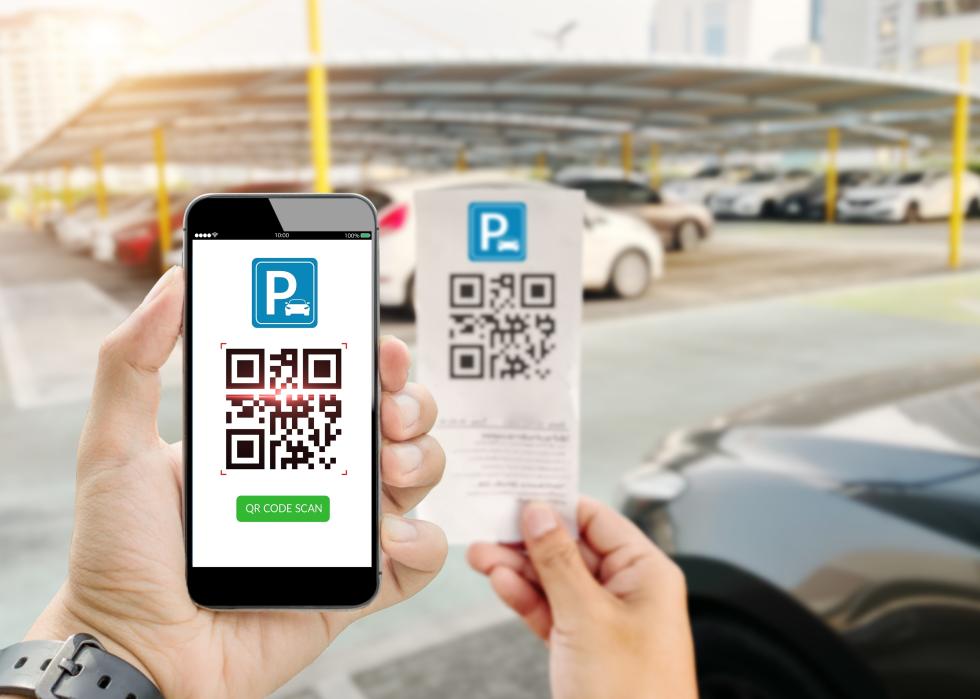 Hand holding phone scanning QR code with blurred parking lot as background.