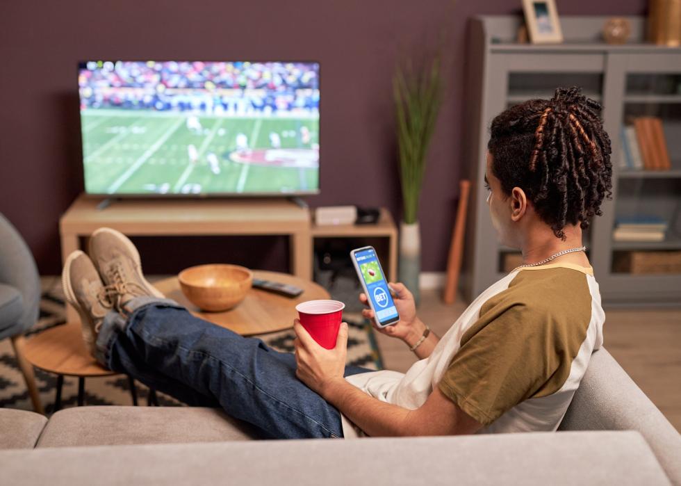 Back view of a young man watching football on couch with online betting app.