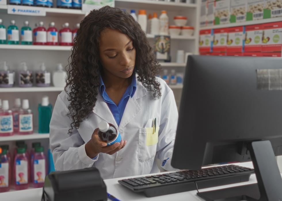 A pharmacist stands in front of a computer in a pharmacy, examining a bottle of medicine.