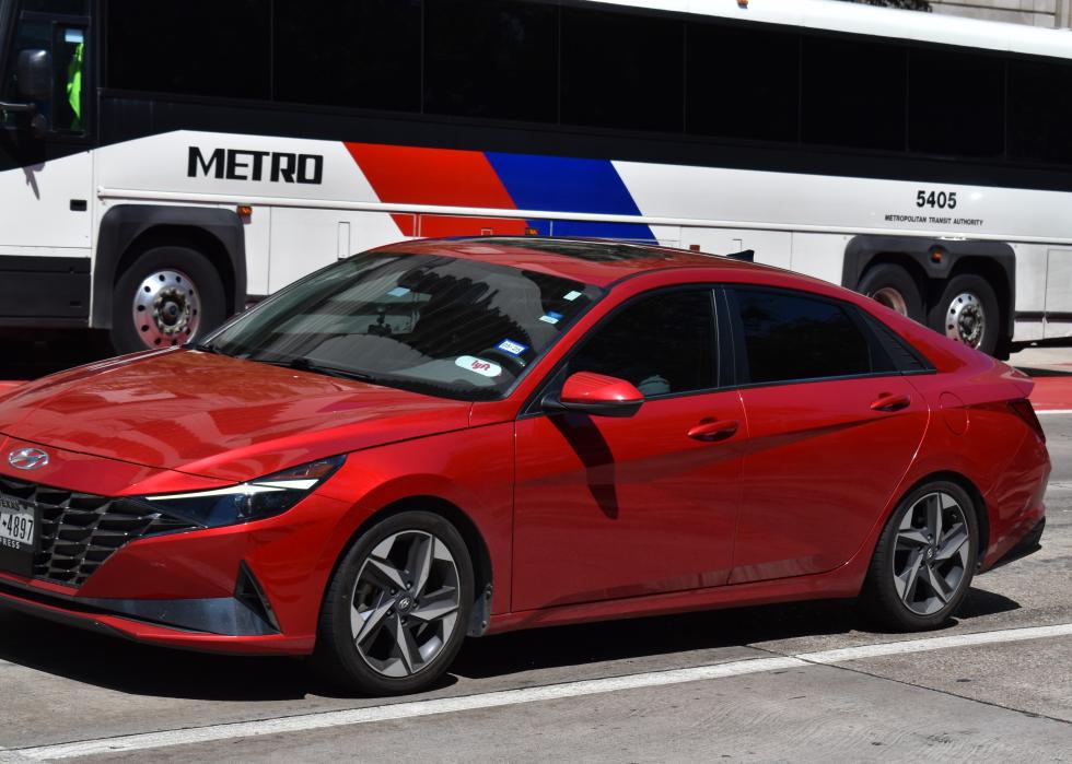 A red Hyundai Elantra Hybrid with a Lyft sticker in the window passing a Metro Bus on the road.