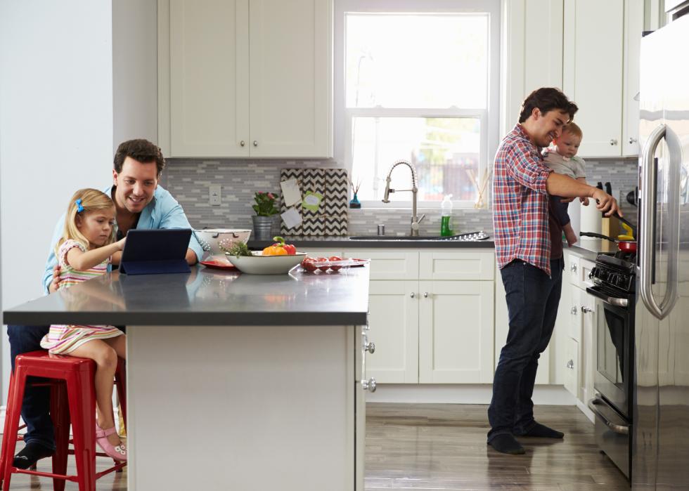 Man playing on tablet with little girl, another man holding baby cooking on stove