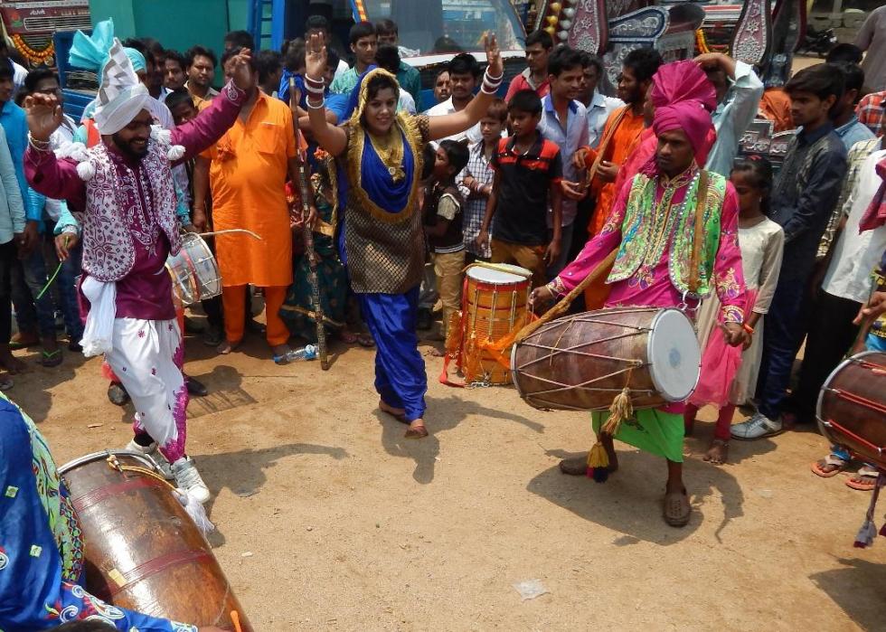 A group of people in colorful outfits dance, sing, and play drums. 
