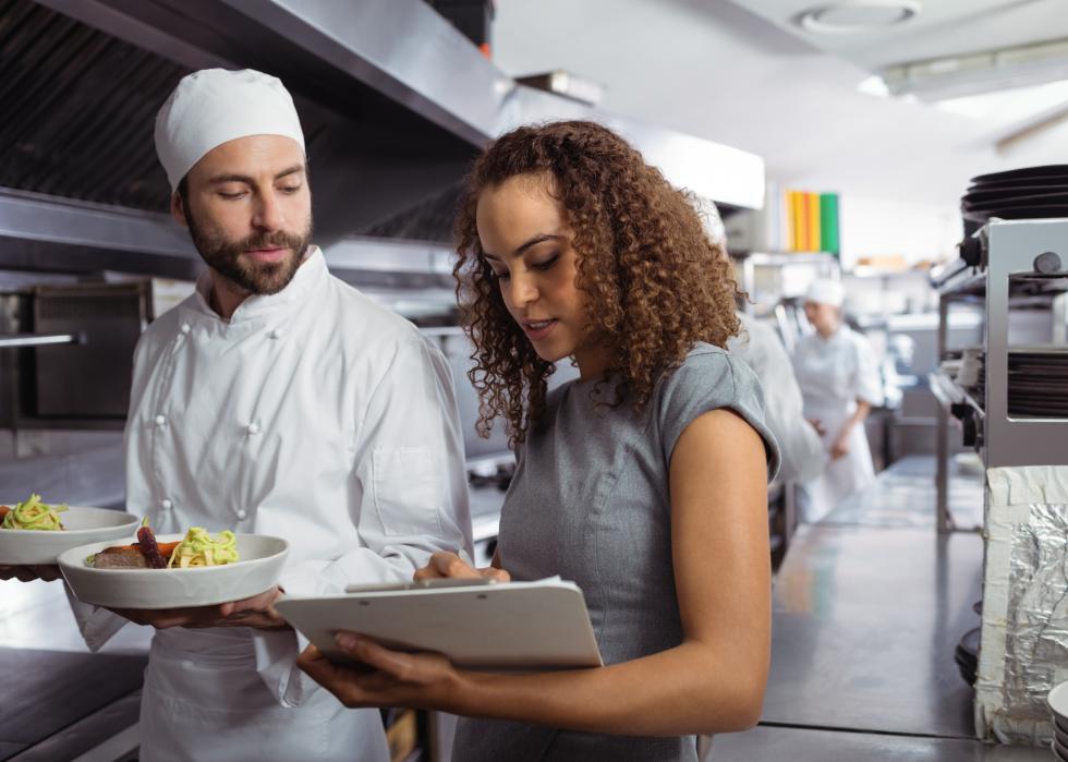 Food service workers discussing menu on a clipboard in a commercial kitchen.