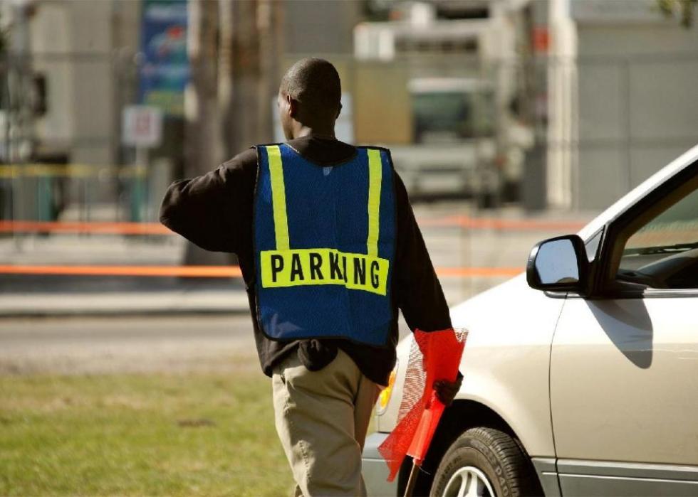 A man wearing a vest with the word parking on it walking outside next to a car.