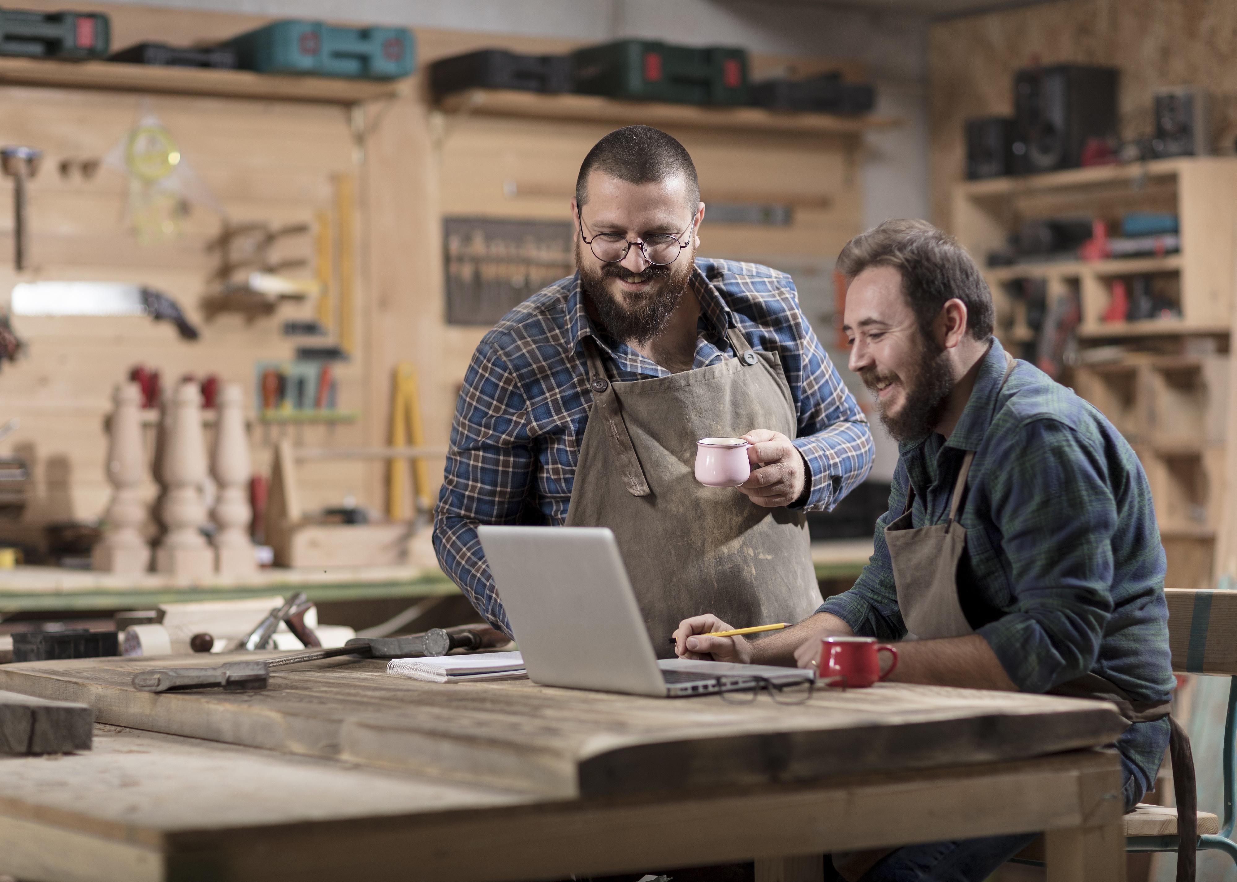 Two wood designers working with laptop in workshop holding small teacups.