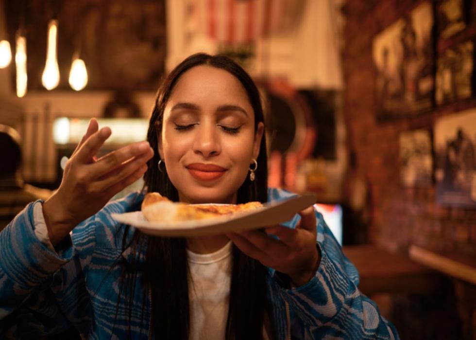 A person smelling the plate of food she is holding up to her face in a dimly lit restaurant. 