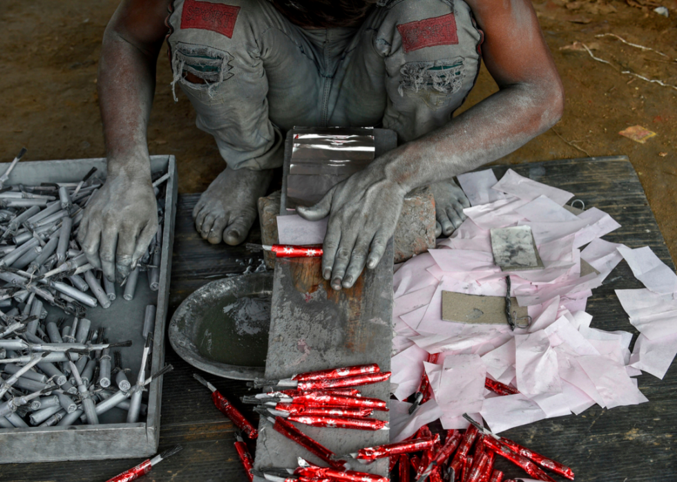 A person covered in gray dust, crouched on the ground, rolling firecrackers in bright red paper. 