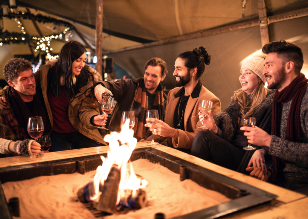 Six people sitting by a fire drinking wine in a covered outdoor area.