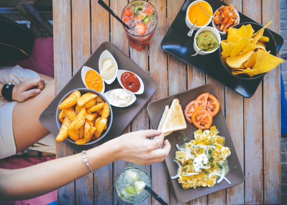 An overhead view of a person sitting at a table with plates of potato wedges, chips, dips, bread, and juice. 