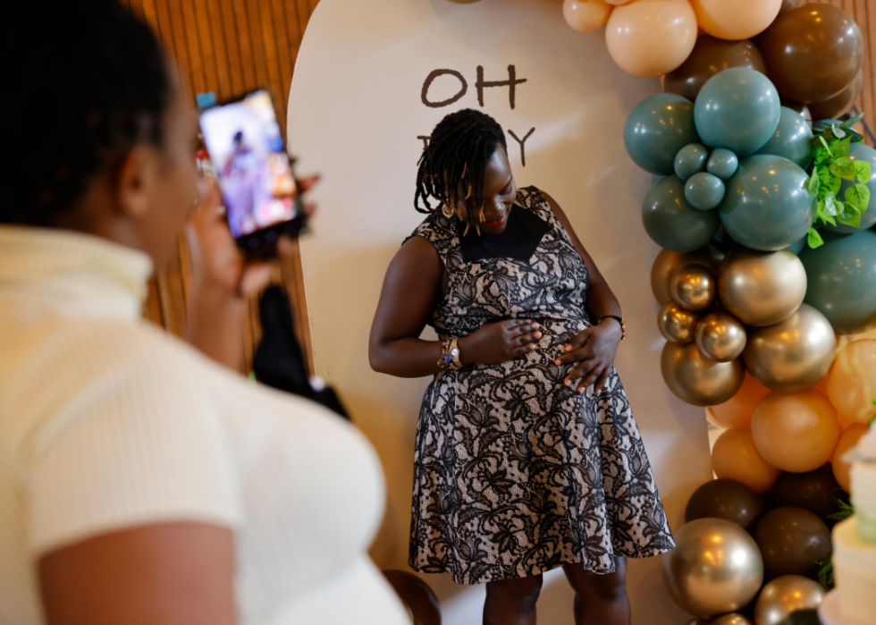 An expecting mother posing for a photo next to turquoise, peach, and gold balloons while someone takes a photo on their smartphone. 