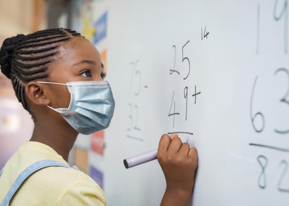 A Black girl wearing a face mask and doing a math equation on a white board at school
