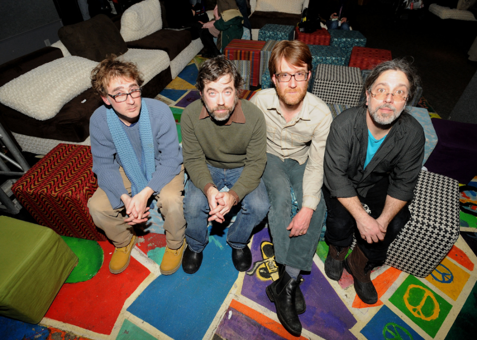 Four bandmembers sit in a very colorful room, looking up toward the camera. 