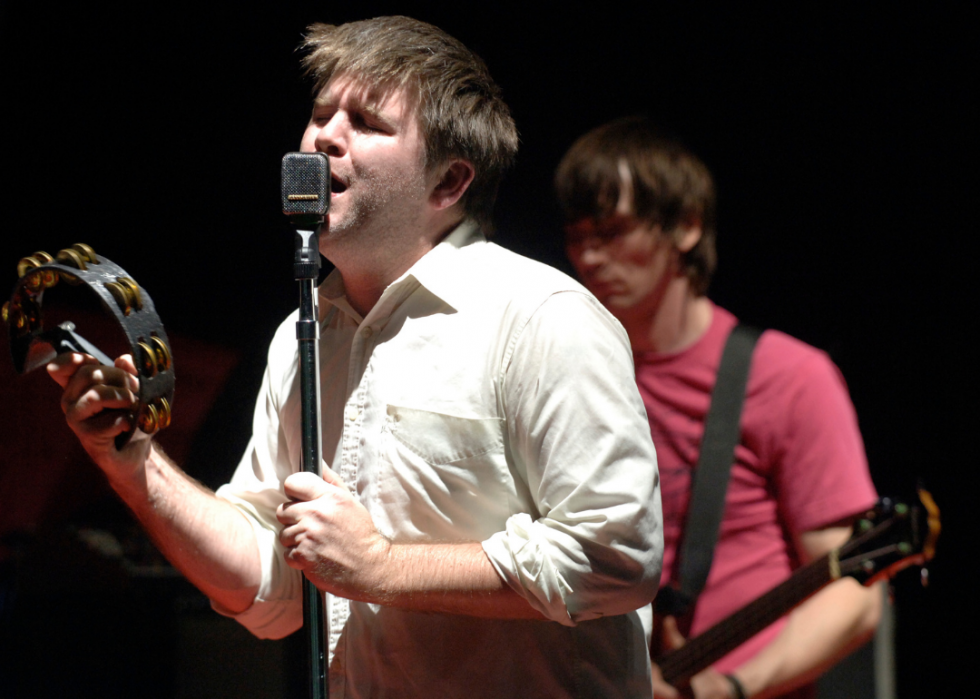 A musician sings into a mic as he plays a tambourine with a guitarist playing behind him.