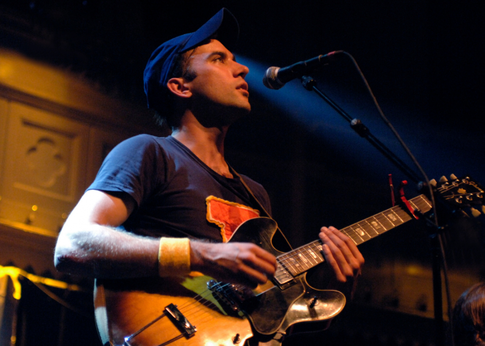 Sufjan Stevens plays guitar while singing into a mic. 
