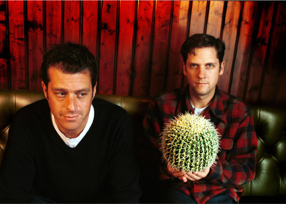 Two band members sit on a sofa, one holding a round cactus. 