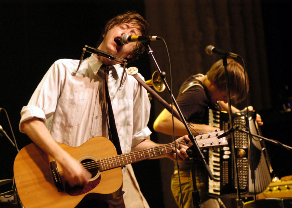 A guitarist sings into a microphone on stage, with an accordion player in the background. 