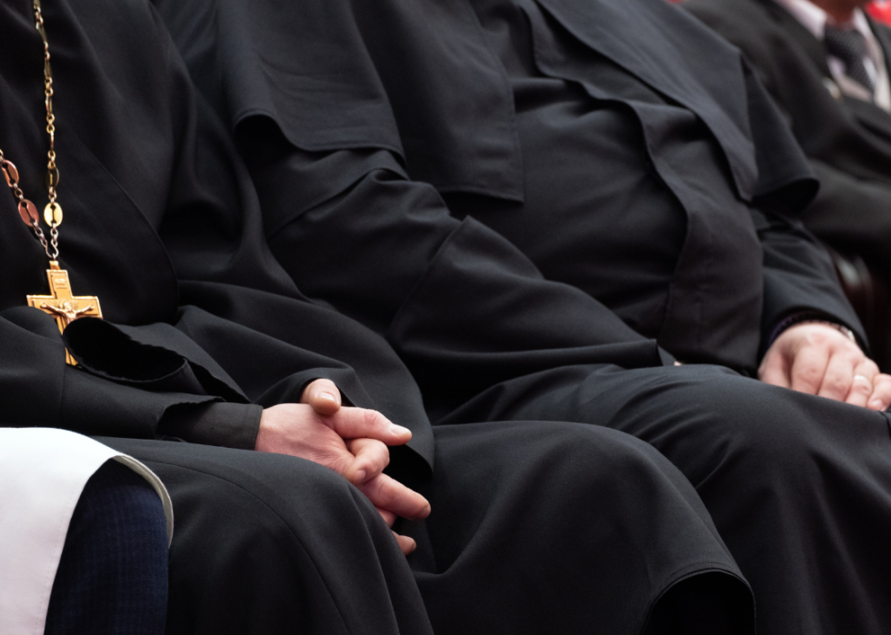 A close up of black-cloaked religious figures sitting in a row, with one cross necklace in view.