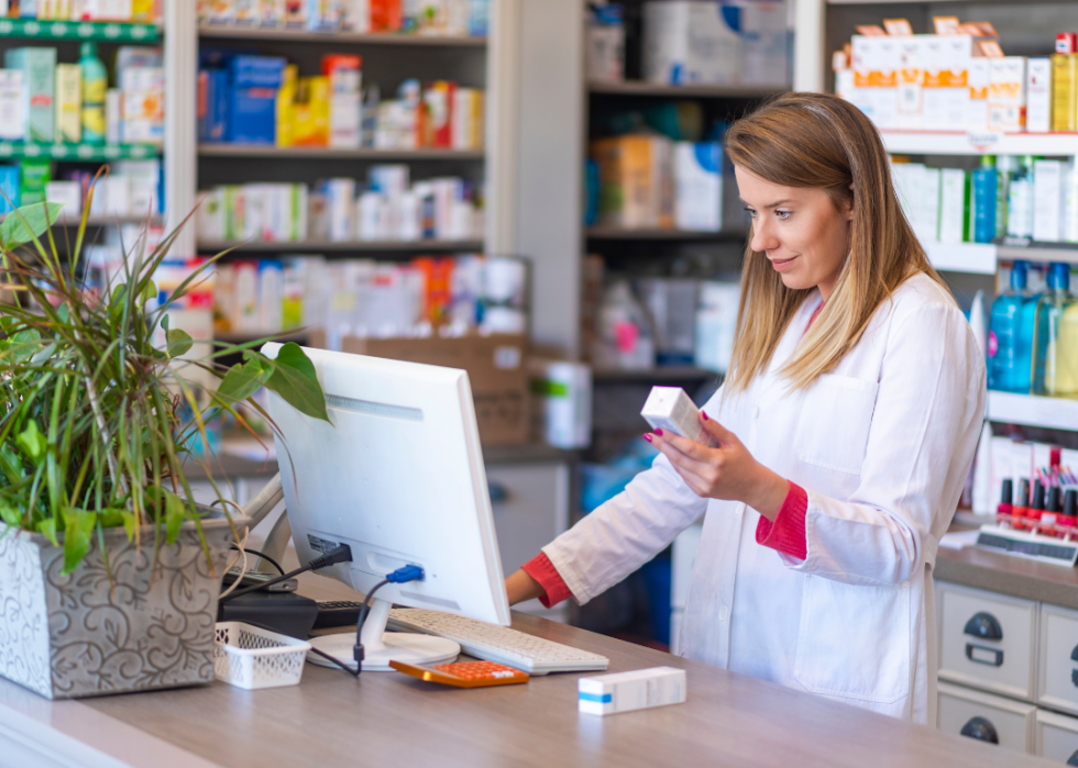 A pharmacist holding a small packaged product as she works on a computer in a pharmacy.