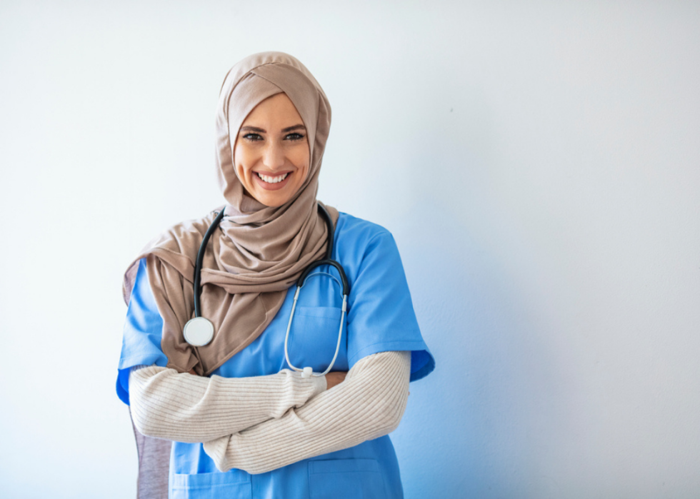 A medical professional in blue scrubs with a stethoscope around her neck, smiling with her arms crossed, posing for a photo.