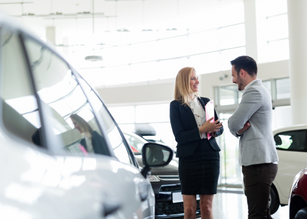 A car salesperson and a customer smile while chatting in a car dealership showroom. 