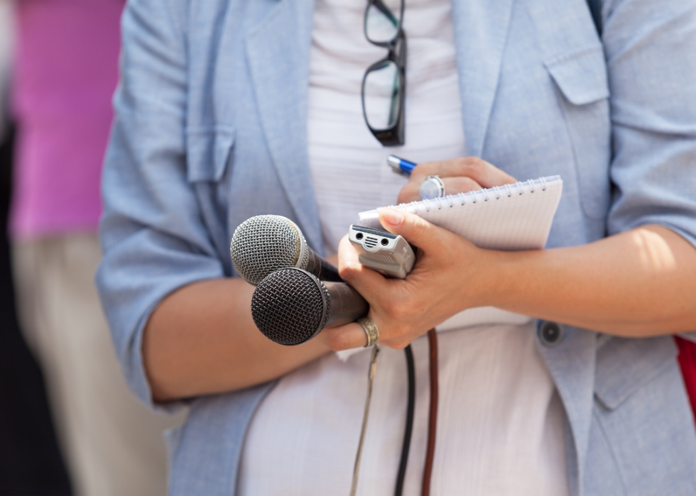 A close up of a pair of hands holding two microphones, a small recording device, and a notebook she is writing in.